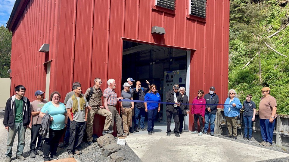 About twenty people stand in front of a red metal powerhouse building as a blue ribbon is cut across its open doorway, with forest on the hillside behind.